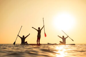 Happy surfers stands on sup boards with raised arms and looks at sunset