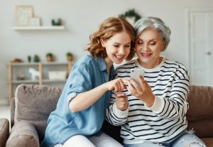 Happy senior woman smiling and sharing data on smartphone with young daughter while sitting on couch at home together