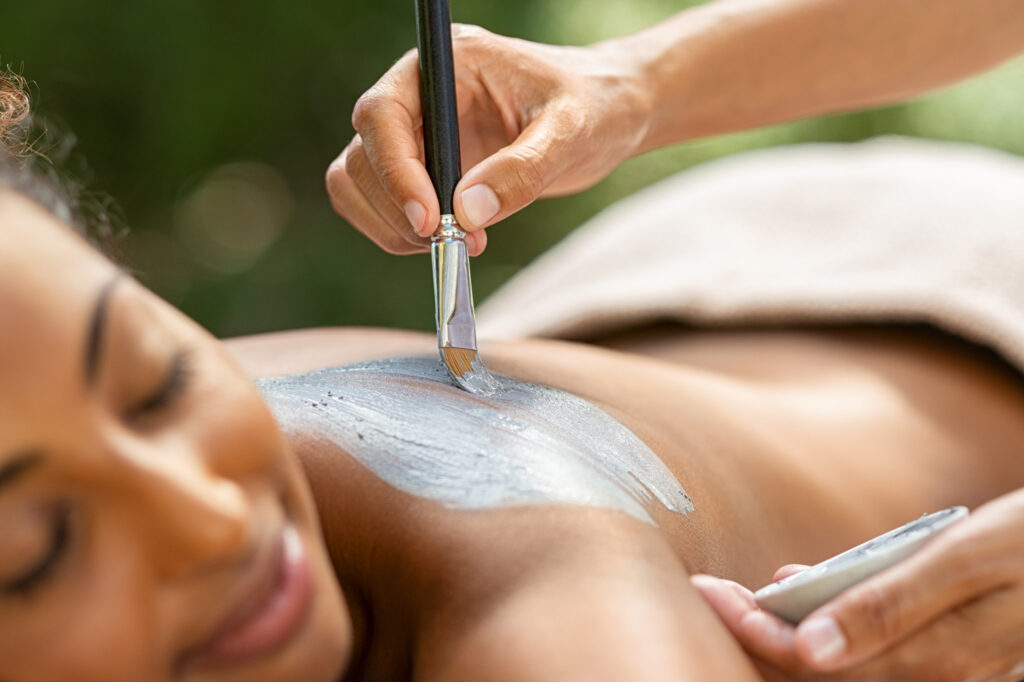 Hands of masseuse applying mud mask on a woman's back. The woman is relaxing with eyes closed while the mask is applied.