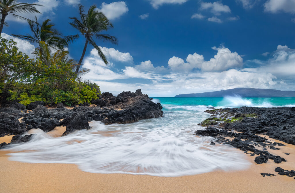 Hawaiian waves crashing onto a rocky shore with a mountainous island in the background.