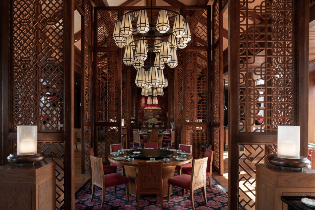 A beautifully decorated private dining room in a traditional Chinese restaurant featuring an ornate wood lattice design, a large round dining table with red cushioned chairs, a central marble lazy Susan, intricate pendant lantern chandelier, and patterned carpet.