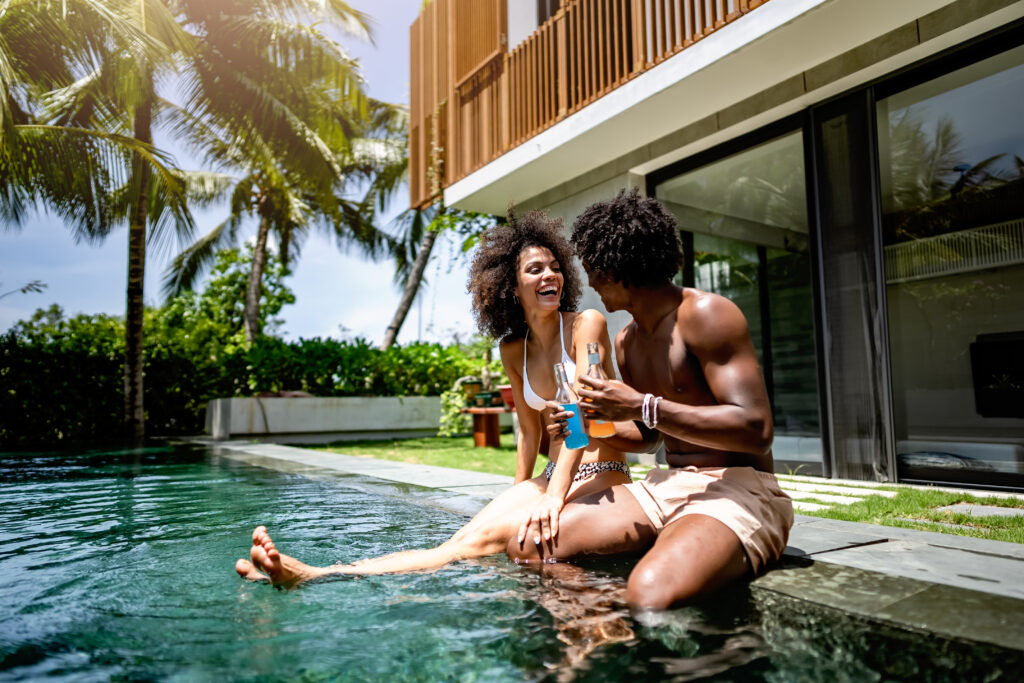 A young couple enjoying a sunny day at a luxurious poolside, sitting at the pool's edge with drinks in hand. 