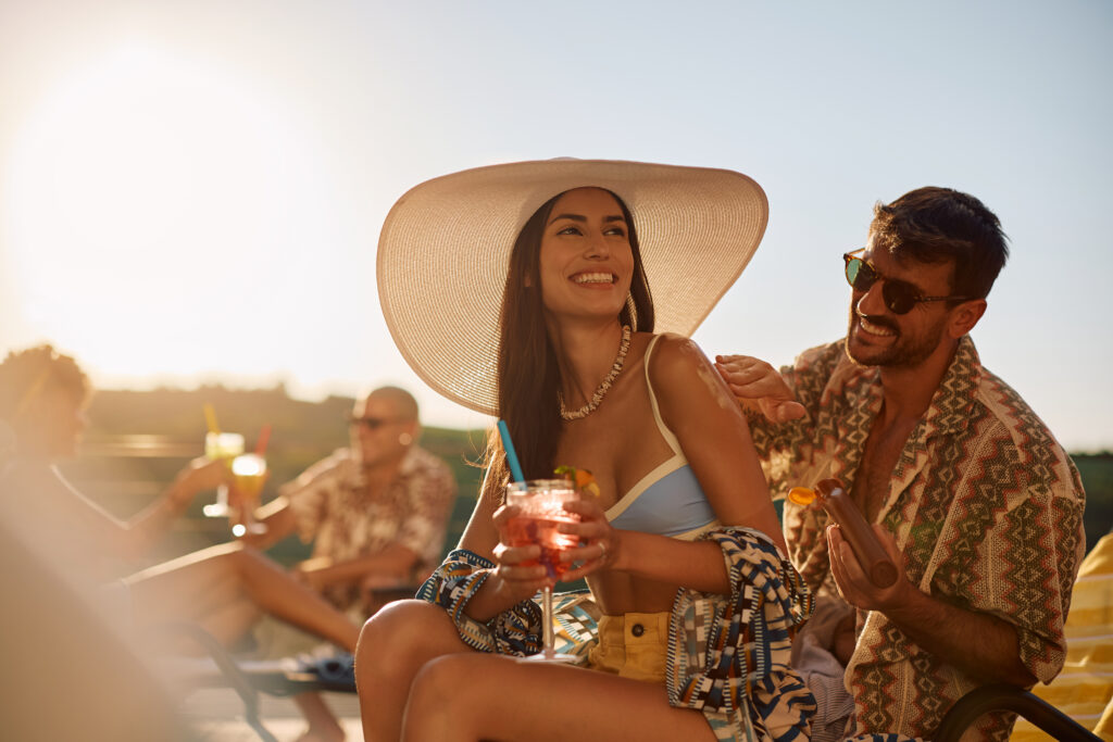 Happy woman enjoying in summer cocktail while her boyfriend is applying suntan lotion on her skin.