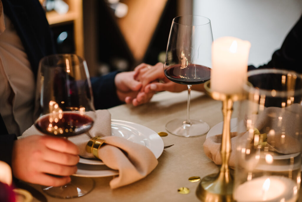 A couple holding hands across a romantic dinner table filled with wine and decorated with gold accents.