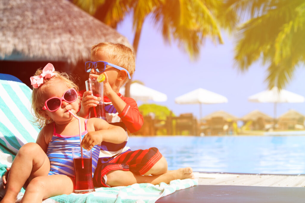 Two young children sit on a towel by a pool, wearing colorful swimsuits and sunglasses, sipping drinks with straws. Palm trees and sun umbrellas are in the background on a bright, sunny day.