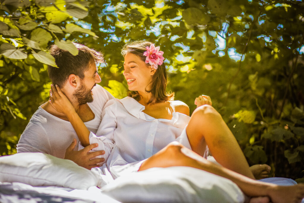 A couple nestled together on a blanket within nature, green tree leaves surrounding them, as they happily smile at one another. 