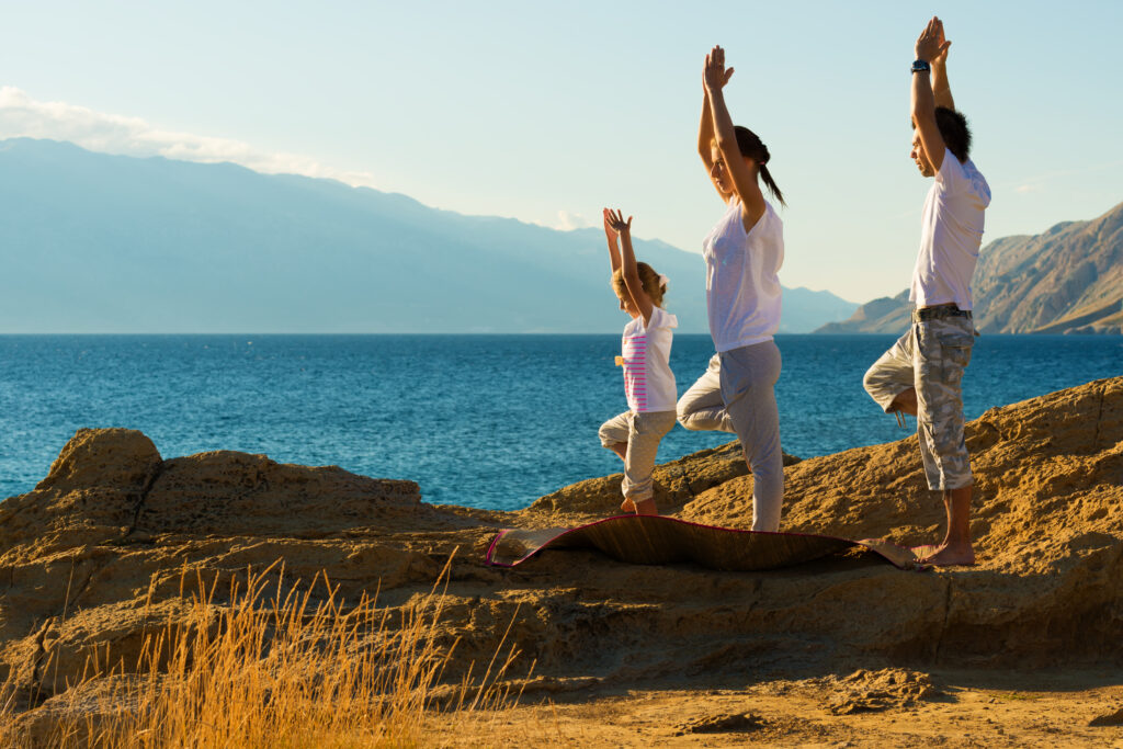 Family on a wellness trip, a 2026 travel trend, doing yoga poses on a rocky beach, staring out at the ocean.