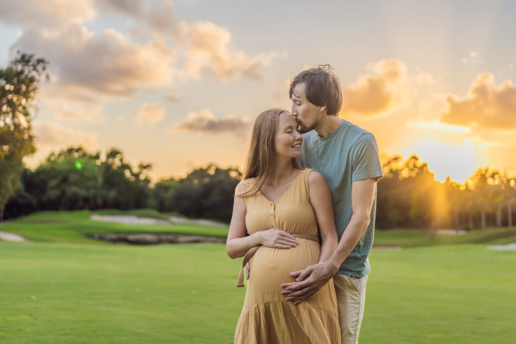 A happy husband and pregnant wife stand together holding their hands over the mother's stomach while standing on a golf course during sunrise on their babymoon getaway.