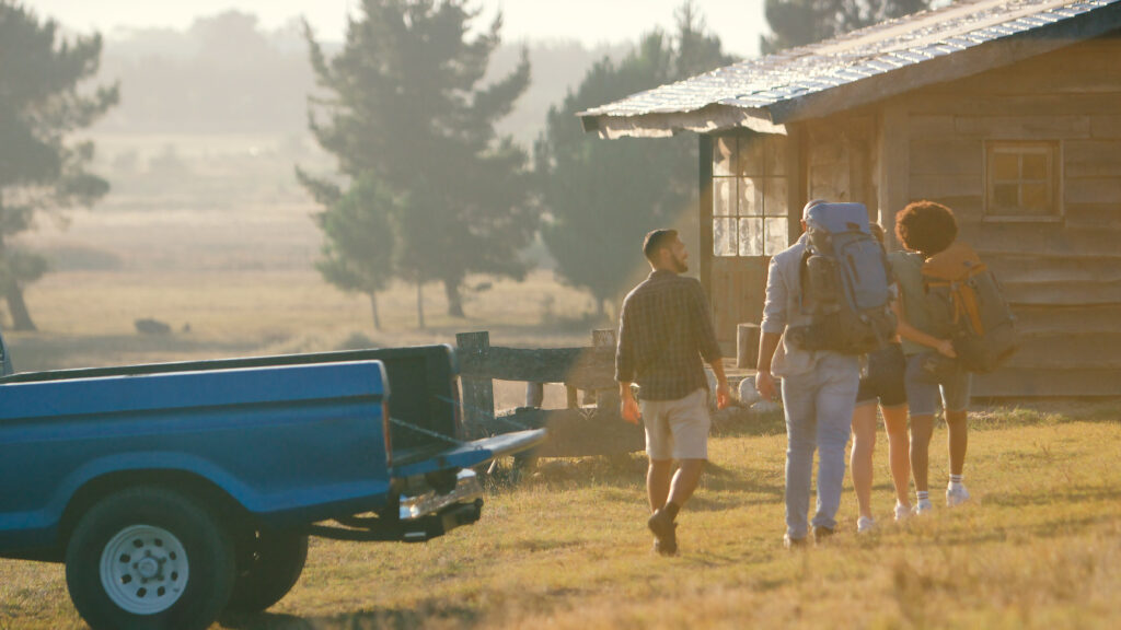 Group Of Friends Unloading Backpack From Pick Up Truck On Road Trip To Cabin In Countryside
