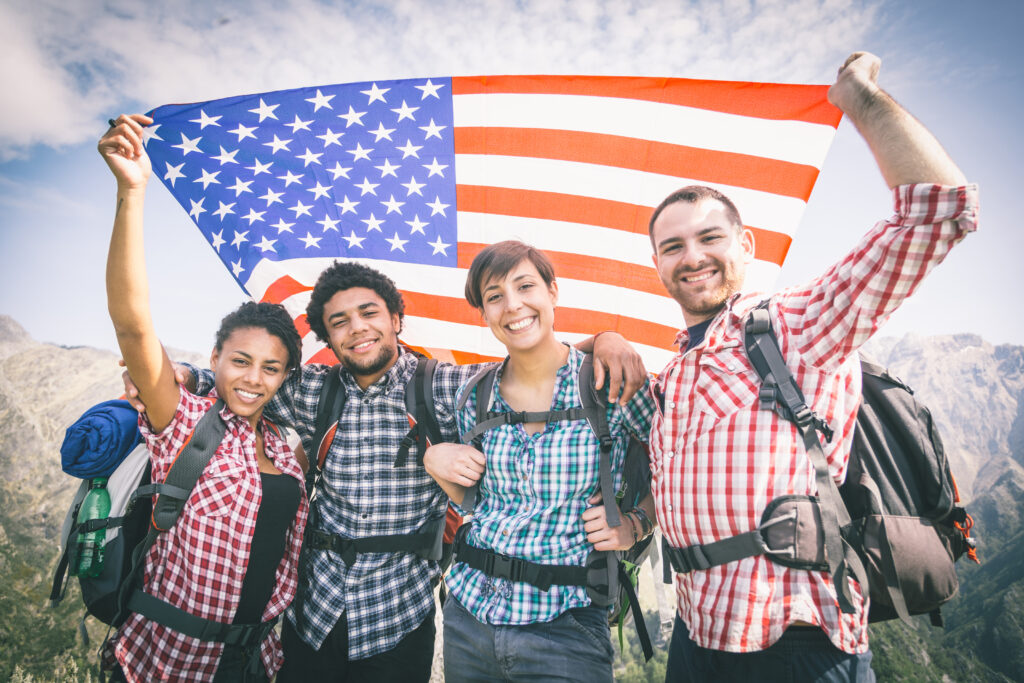 Four people holding a large American Flag above their heads while standing on top of a mountain. 
