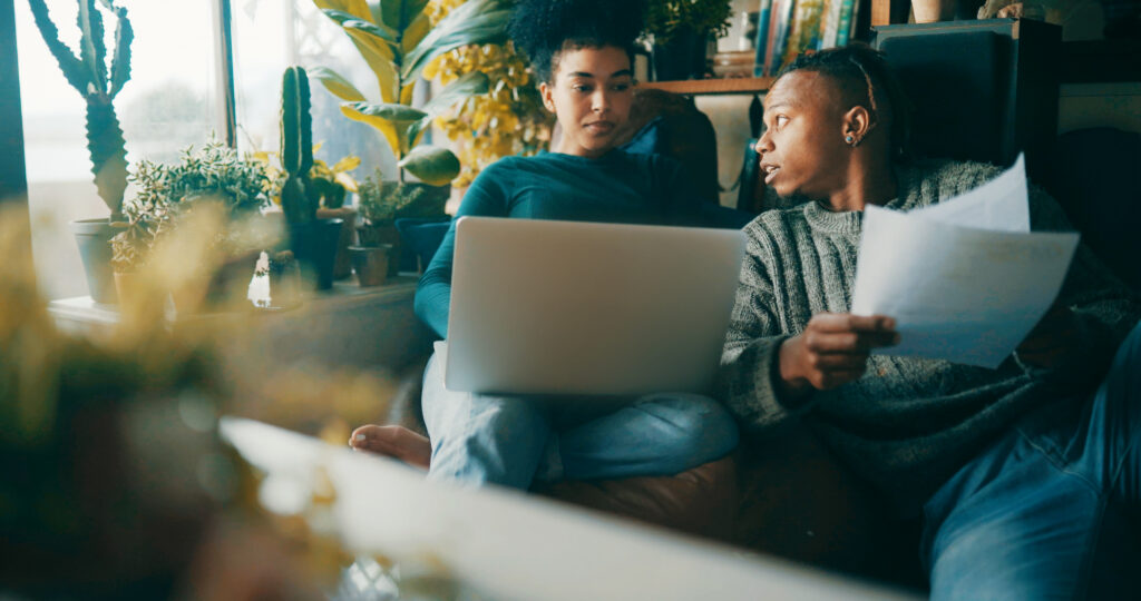 Two people lounging on a couch while one reviews documents and the other views a computer.