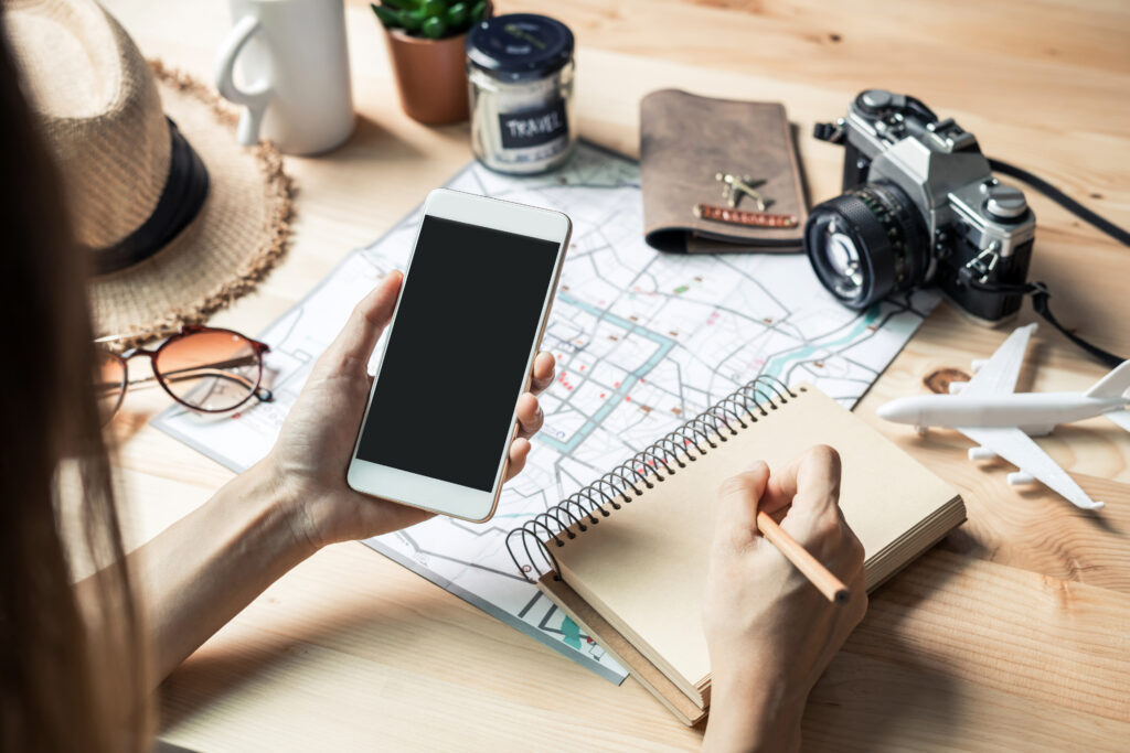 A camera, map and other travel essentials on a table while a women reviews her phone.