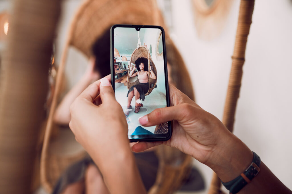 A close-up of someone taking a picture of someone else as they sit in a hanging wicker chair at a small venue.