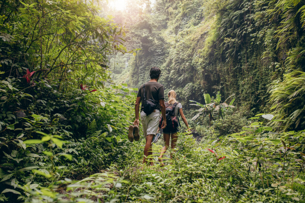 A man and woman walking barefoot through a dense jungle, surrounded by trees and flora.