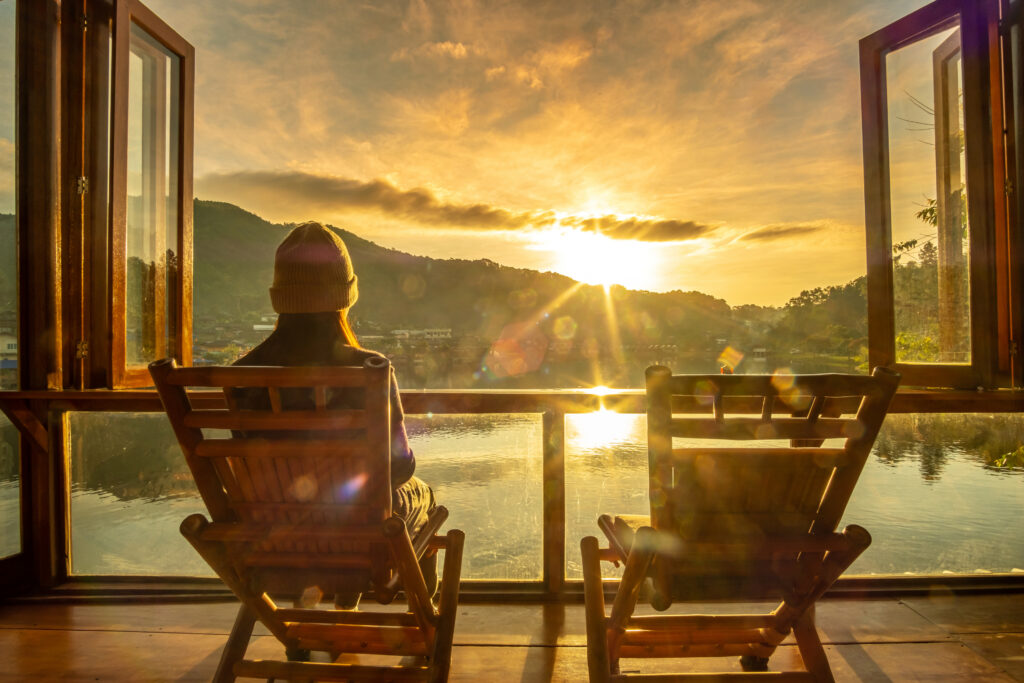 A woman wearing a beanie, staring out into a lake with a mountainous expanse in front of her while the sun sets, a perfect activity when looking to travel with purpose.