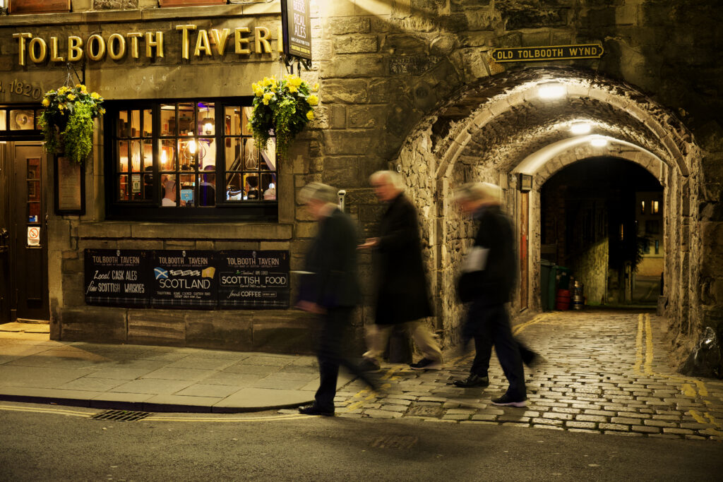 A group of men walking down an old street near Old Tolbooth Wynd, in Scotland, their bodies blurry from the moving camera.
