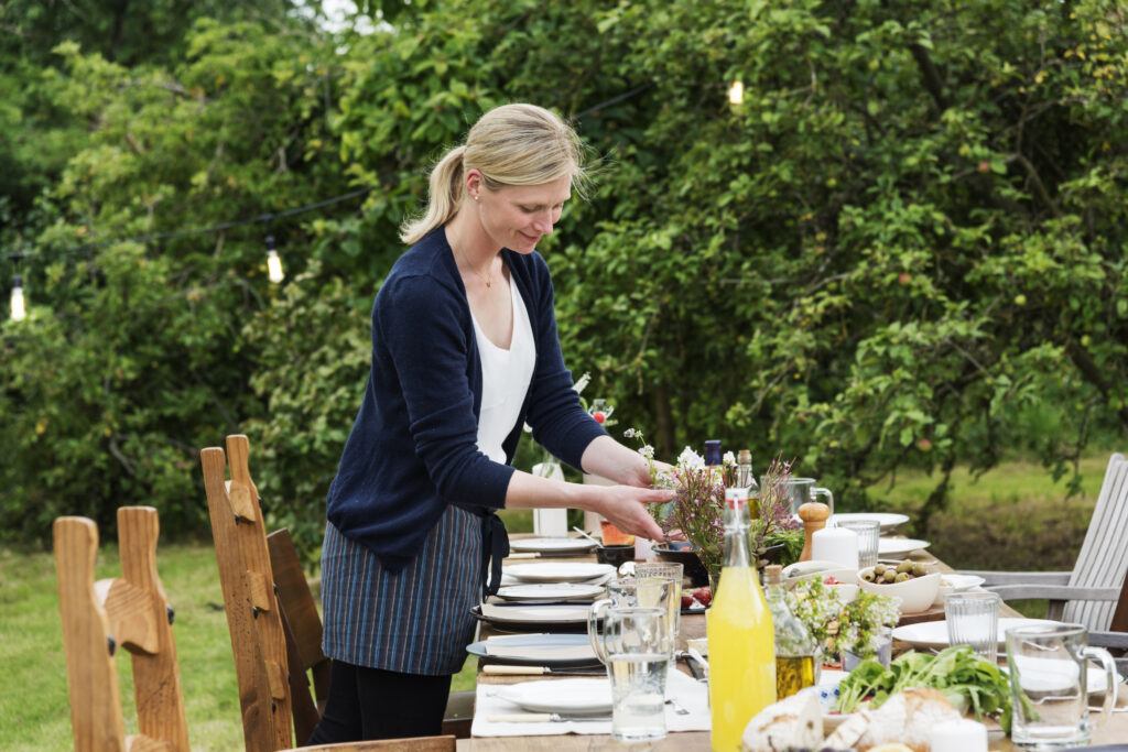 A women standing in front of a long banquet table surrounded by dining chairs and settled within an orchard.
