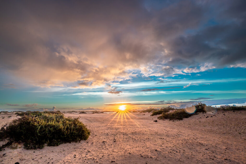 A stunning view of a sun setting in the background of a sandy beach with the cloudy sky shifting between day and night.