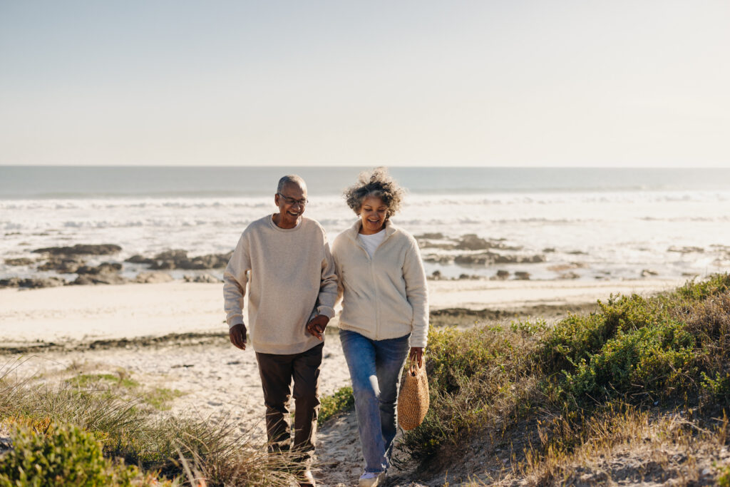 A happy older couple, smiling while holding hands and walking through the sand on the beach during their quietcation. 