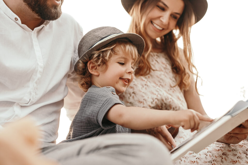 Two parents and a child sitting outside together happily, reviewing a book, with the son pointing to the contents within it.