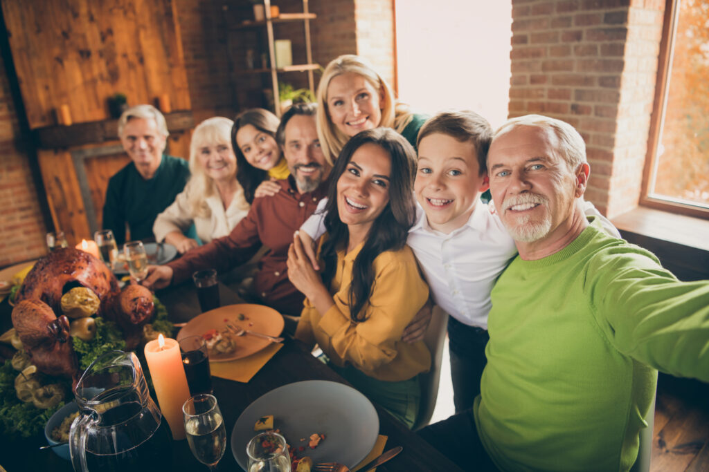 A happy family across generations standing behind a dinner table with a large turkey on top of it, smiling at the camera.