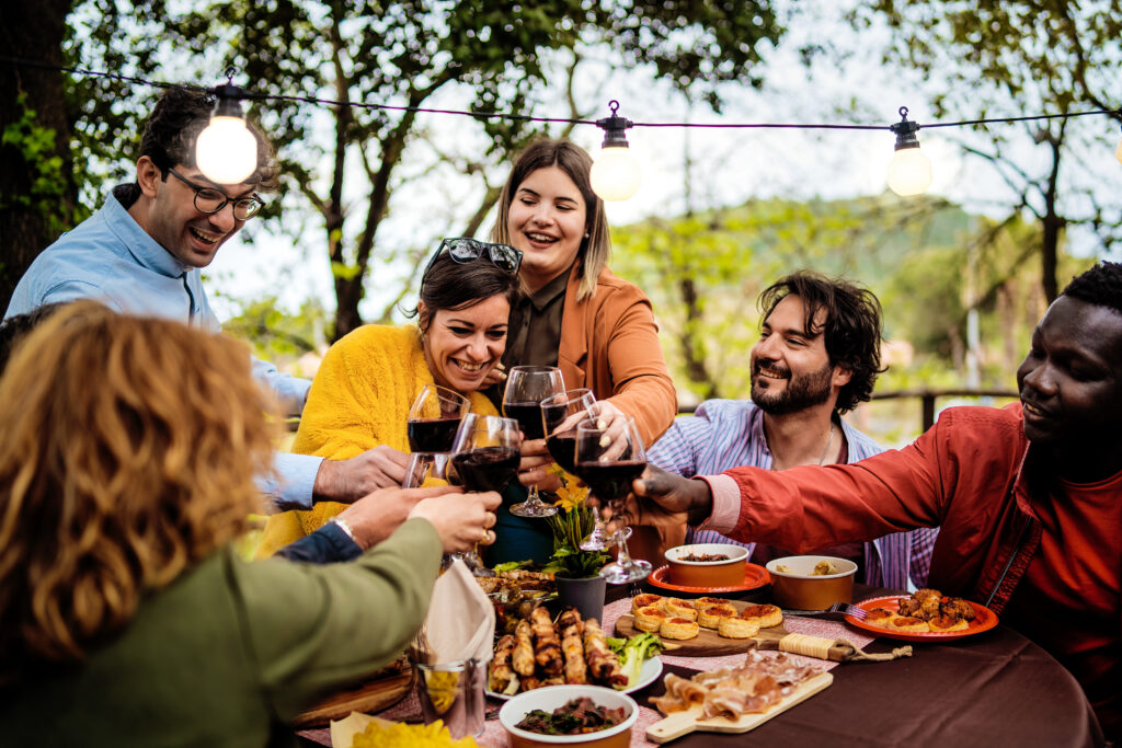 A group of friends sitting around a table filled with plated foods, smiling and toasting with glasses of wine during their leisure travel.