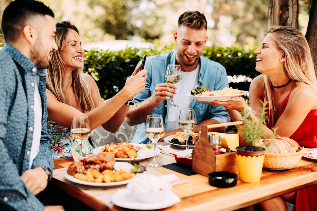 Four friends sitting together at a dinner table, taking photos of their aesthetic food, supporting leisure travel trends.
